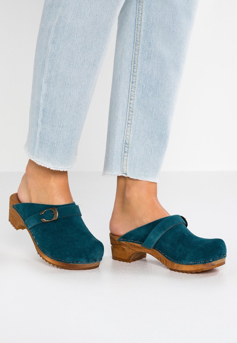 Pieds portant des sabots en daim bleu sarcelle avec des semelles en bois et une bride à boucle, associés à un jean en denim bleu clair aux ourlets effilochés.