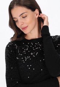 Fekete pulóver, a felső részén flitterekkel díszítve, bordázott mandzsettákkal és kerek nyakkivágással. Sima anyag csillogó részletekkel.