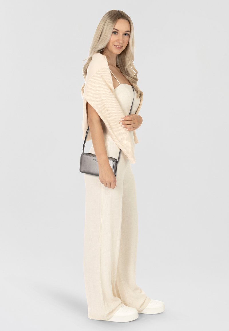 Beżowy żakiet ze żłobieniami na dopasowanym, jasnobeżowym kombinezonie, z wąską czarną torebką na ramię i białymi sneakersami. Modelka stoi bokiem.
