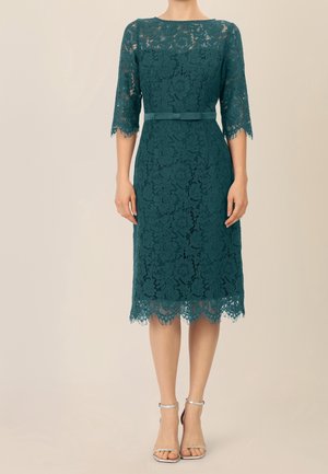 Robe en dentelle couleur sarcelle à longueur genou, manches trois-quarts, taille cintrée avec ceinture à nœud, et ourlet festonné, portée avec des talons argentés à bride cheville.