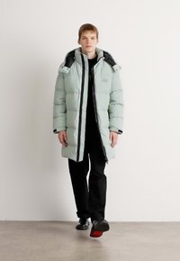 Svijetlozelena puffer jakna s kapuljačom, s crnim oblogama i zatvaračem. Model nosi crne hlače i cipele. Glatka, podstavljena tekstura.