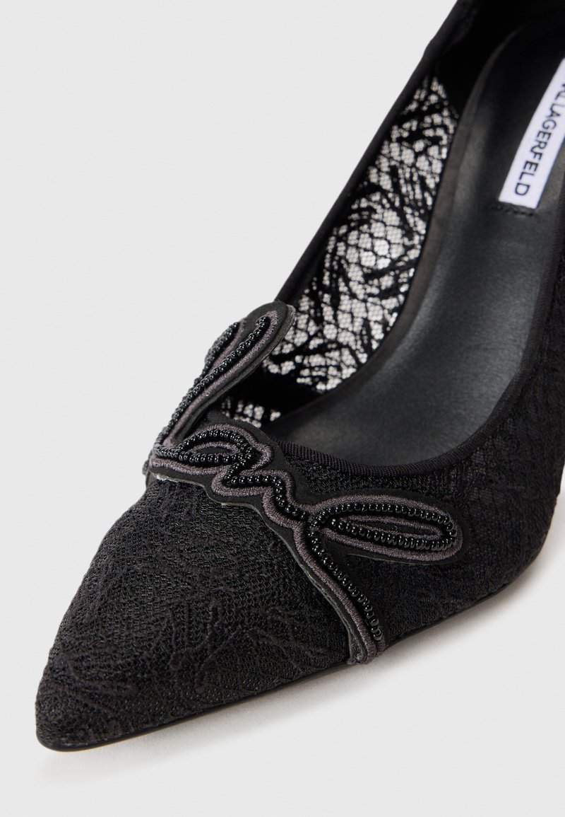 Schwarze Spitzenpumps mit spitzem Fuß, ausgestattet mit einem kunstvollen Stoffmuster und dekorativen Perlenakzenten entlang des oberen Randes.