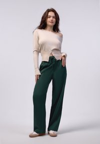 Bežna skrajšana pletena jopica z detajli z gumbi, v kombinaciji s temno zelenimi širokimi hlačami. Model nosi bež natikače. Gladka tekstura po celi površini.