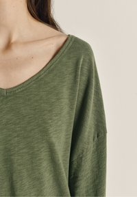 Zaļa pārāk liela v veida tshirts, kas izgatavota no mīksta, teksturēta auduma. Piedāvā brīvu piegriezumu ar nolaistām plecu šuvēm un vienkāršu, neizpušķotu dizainu.