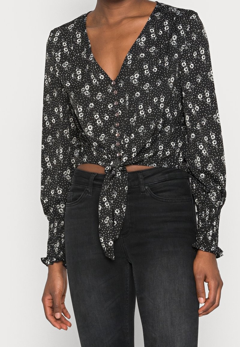 Blusa nera a fiori con maniche lunghe, davanti con nodo, scollo a V e dettaglio dei bottoni. Presenta motivi floreali bianchi e sottili pois sul tessuto.