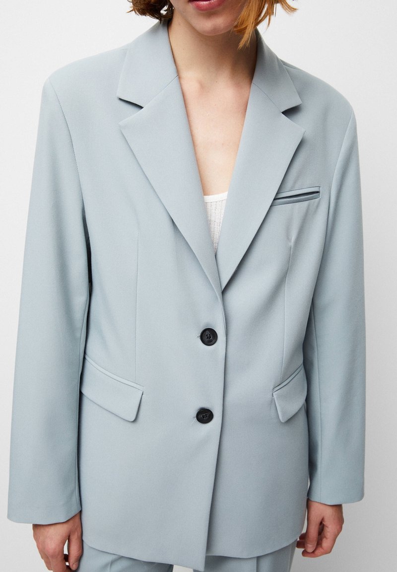 Femme portant un blazer bleu clair à double boutonnage avec revers crantés et deux poches avant à rabat, sur un haut blanc, devant un fond uni.