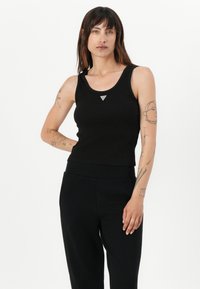 Svart ribbet tanktop med rund hals, med en liten trekantet sølvdetalj på brystet. Modellen har på seg svarte bukser.