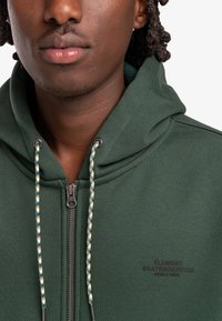 Grøn zip-up hættejakke med tekstureret finish, med en kontrasterende snor til hætten og trykt logo på brystet.