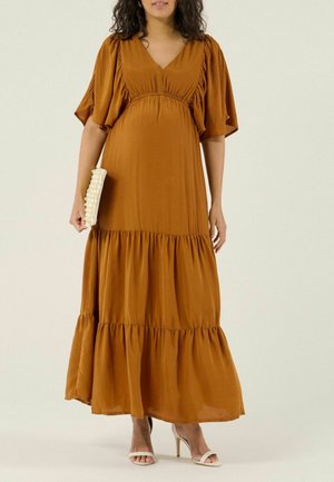 Vrouw die een roestkleurige maxi-jurk met laagjes en wijd uitlopende mouwen draagt, een crème gevlochten clutch vasthoudt en witte sandalen met hoge hakken draagt.