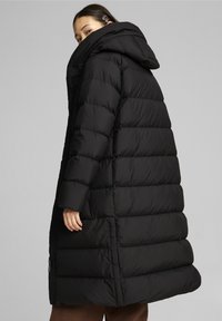 Longue doudoune noire avec capuche, comportant de larges sections matelassées et une coupe légèrement oversize. Fermeture éclair et poches latérales visibles.