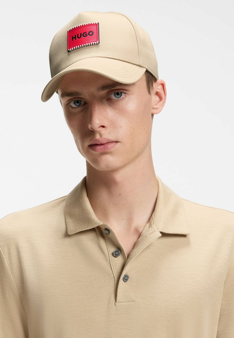 Jeune homme portant une casquette beige avec un écusson logo HUGO rouge et un polo beige assorti sur un fond uni.