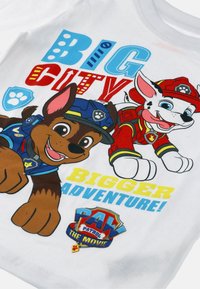 Biała bawełniana koszulka z kolorowymi postaciami psów z kreskówek w niebieskich i czerwonych mundurach. Tekst brzmi "WIELKIE MIASTO, JESZCZE WIĘKSZA PRZYGODA!" oraz "PAW PATROL: FILM."