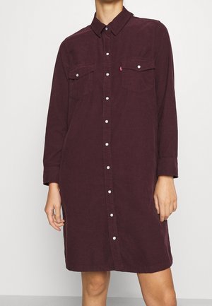 Mujer que lleva un vestido camisa de pana de manga larga, abotonado, de color burdeos oscuro, con dos bolsillos en el pecho y botones de presión blancos.