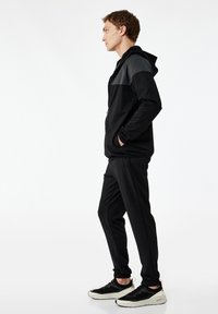 Czarny sportowy dres z zamkiem błyskawicznym i szarym panelem w bluzie z kapturem, slim-fit spodnie oraz strukturalne czarne sneakersy. Gładki materiał i minimalistyczny design.