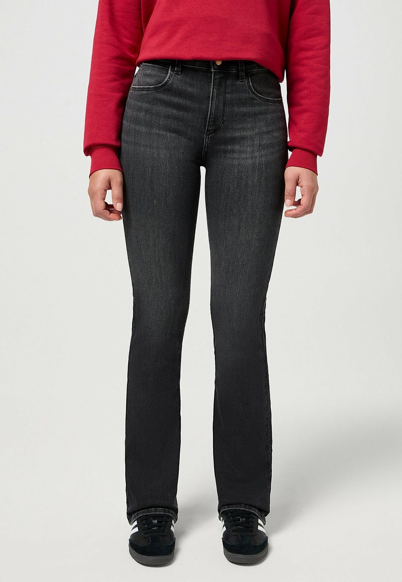 Personne portant un jean slim noir, des baskets noires avec des bandes blanches et un haut rouge à manches longues, debout devant un fond uni.