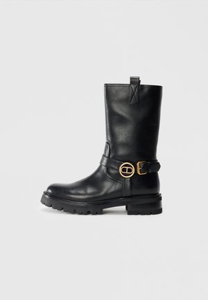 Black mid-calf leather boot with chunky sole, decorative strap, and gold-tone round and rectangular buckles, shown from the side on plain background.