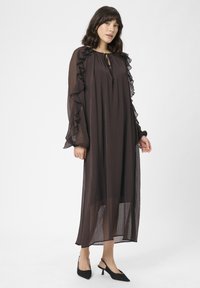 Žena v dlhých, priehľadných tmavo hnedých šatách s volánovými rukávmi a čiernymi slingback lodičkami, postavená pred jednoduchým bielym pozadím.