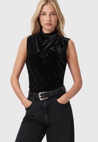 Blonde vrouw in een mouwloos zwart fluwelen top en zwarte jeans met een zwarte riem, met handen in de zakken, neutrale grijze achtergrond.