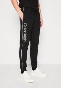 Black sweatpants with white side stripes, drawstring waist, and embroidered "Calvin Klein" logo, paired with white sneakers.