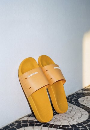 Paire de sandales slides en caoutchouc jaune avec semelles texturées reposant contre un mur blanc sur un sol en mosaïque noir et blanc.