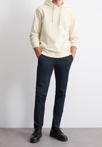 Felpa beige con cordino, abbinata a pantaloni blu scuro. Le scarpe nere hanno una suola spessa. Texture dei tessuti liscia in tutto.