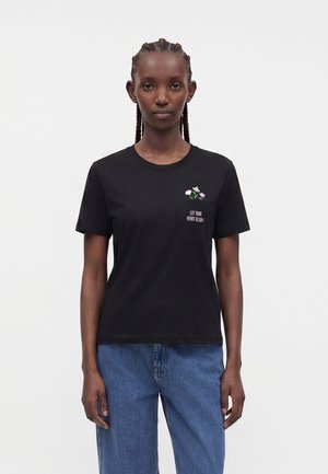 Mujer con cabello trenzado que lleva una camiseta negra con un pequeño bolsillo, diseño floral y texto "Deja que tu corazón florezca" combinada con vaqueros azules.