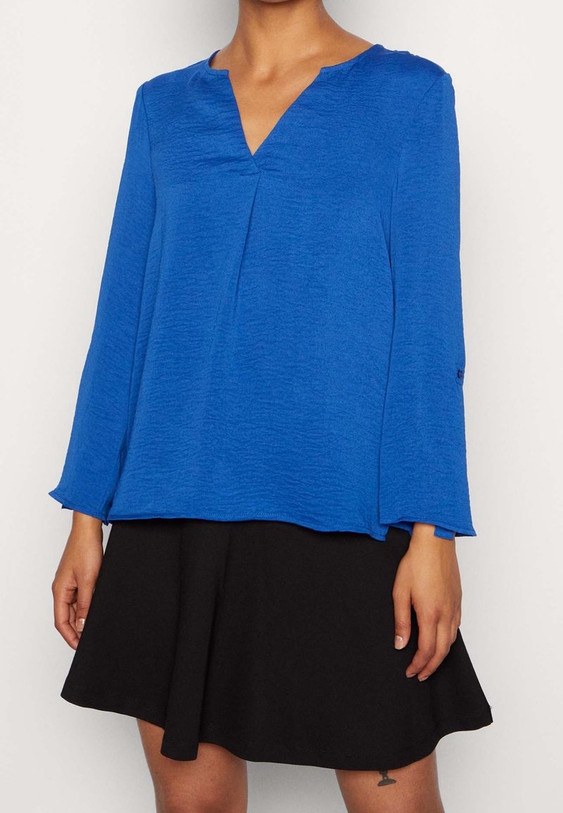 Kobieta ubrana w bluzkę z długim rękawem w kolorze kobaltowym z dekoltem w kształcie litery V i czarną, rozkloszowaną spódnicę na jednolitym tle.