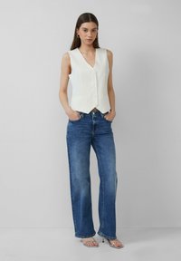 Weißer strukturierter Weste mit tiefem V-Ausschnitt und fünf Knöpfen, kombiniert mit hochgeschnittenen blauen weiten Jeans. Schlichter Hintergrund, keine zusätzlichen Details.