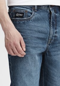 Vidēji zila džinsa bikses ar klasisku taisnu griezumu. Aprīkotas ar priekšējo kabatas etiķeti un šuvju akcentiem. Materiāls izskatās mīksts ar vieglu tekstūru.