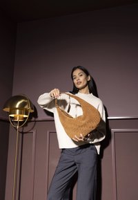 Gewebte, tanfarbene Leder-Schultertasche mit einer geschwungenen Silhouette und einem breiten Tragegurt. Das Modell trägt einen cremefarbenen Pullover und graue Hosen vor einer dunklen Wand.