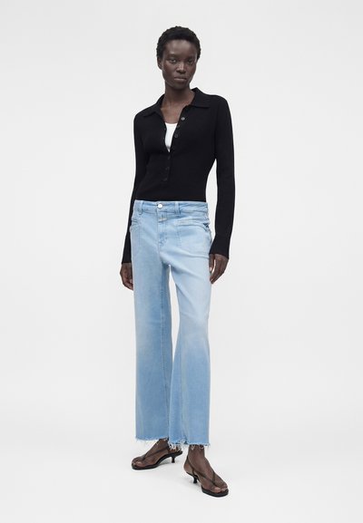 Γυναίκα που φοράει μαύρο ριμπ πουλόβερ με κουμπιά, λευκό τοπ από μέσα, ανοιχτό μπλε παντελόνι με φαρδιά πόδια και ακατέργασμα τελειώματα, μαζί με μαύρα σανδάλια με τακούνι.