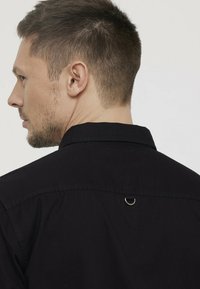 Zwarte shirt met een kraag, met een metalen lusdetail op de achterkant. De textuur is glad met een lichte glans.