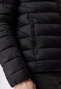 Giubbotto a sacco nero con sezioni a cerniera orizzontali, una tasca laterale con zip e polsini elasticizzati per un maggiore calore e comfort.