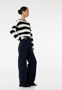 Gestreepte marineblauwe en witte cardigan met knopen, gecombineerd met wijde marineblauwe broeken en puntige zwarte schoenen. Glad textuur en gestructureerde pasvorm.