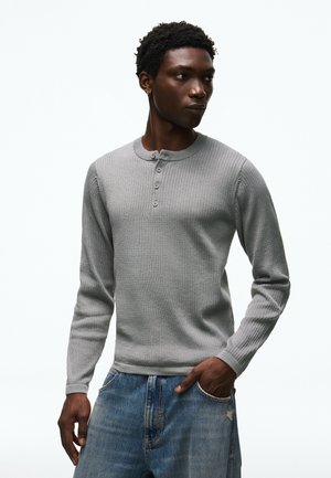 Mann trägt ein hellgraues, geripptes Langarm-Henley-Shirt und blaue Jeans, schaut zur Seite mit einer Hand in der Tasche.