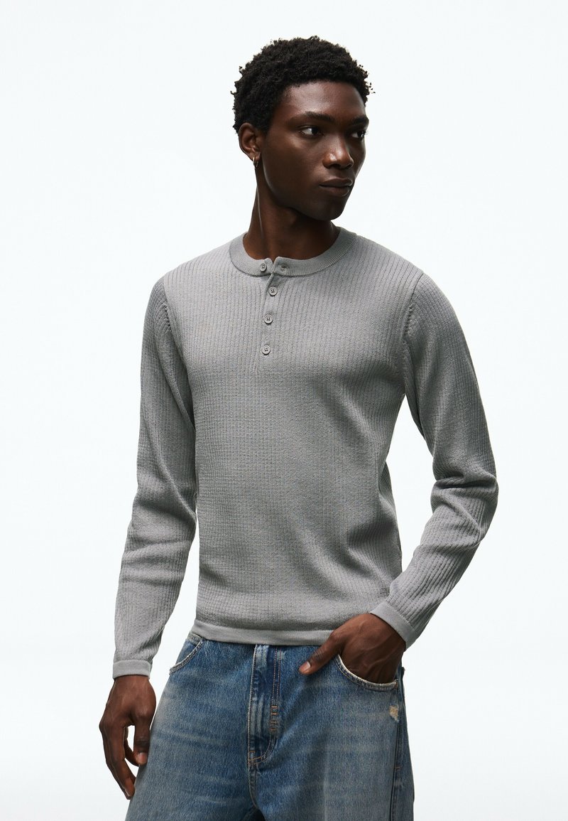 Homme portant une chemise henley à manches longues côtelée gris clair et un jean bleu, regardant sur le côté avec une main dans sa poche.