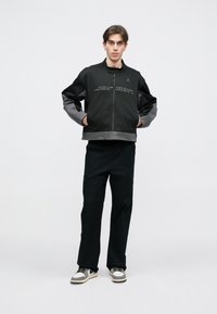 Casaco de fecho éclair em preto e cinza com o logótipo "Jordan" na frente, apresentando um colarinho alto e um corte largo, combinado com calças e sapatos pretos.