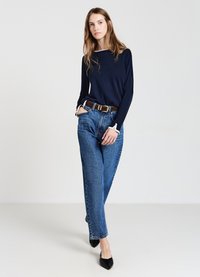 Marineblaues Langarmoberteil mit weißem Saum, kombiniert mit hoch-taillierten blauen Jeans und schwarzen spitzen Schuhen. Das Model steht mit einer Hand in der Tasche.