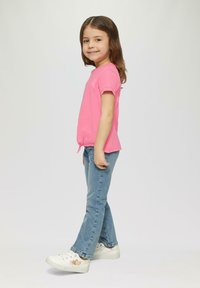 Rosa T-Shirt mit kurzen Ärmeln und Schleifendetail, kombiniert mit hellblauen Jeans und weißen Sneakers, die bunte Herz- und Sternmuster aufweisen.