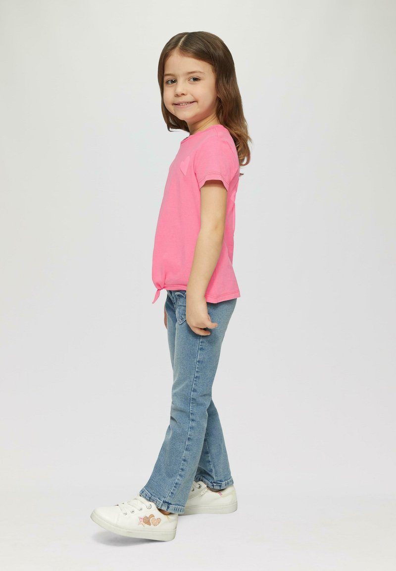 Rosa T-Shirt mit kurzen Ärmeln und Schleifendetail, kombiniert mit hellblauen Jeans und weißen Sneakers, die bunte Herz- und Sternmuster aufweisen.