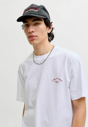 Hvid T-shirt med korte ærmer, med rødt broderet tekst. Båret med en sort kasket med buet skygge og tekstureret stof. Sølv kæde halskæde.