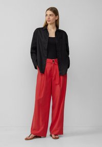 Schwarzes Leinenhemd, schwarzes Top, rote weit geschnittene Hose mit Gürtel und Schnallen-Detail, kombiniert mit braunen Sandalen. Modell steht vor einem neutralen Hintergrund.
