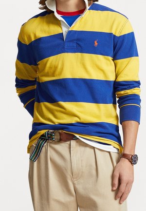 Osoba ubrana w niebiesko-żółtą koszulę rugby w paski, beżowe spodnie z paskiem w paski oraz brązowy skórzany zegarek na nadgarstku.