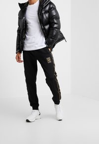 Černá prošívaná bunda s lesklým povrchem, bílé tričko, černé tepláky se zlatými bočními pruhy a logem, doplněné bílými teniskami.