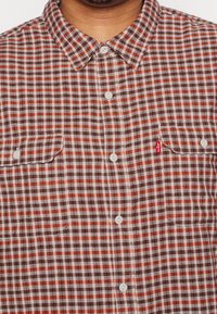 Camisa de franela a cuadros en rojo, marrón y blanco con cuello, dos bolsillos frontales y botones blancos. Cuenta con una pequeña etiqueta de marca en el bolsillo.