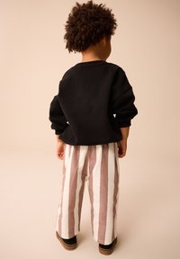 Bambino con capelli ricci che indossa una maglia nera a maniche lunghe, pantaloni larghi a righe e scarpe marroni, in piedi di spalle su uno sfondo neutro.