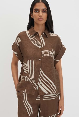 Femme aux longs cheveux foncés portant une chemise marron à manches courtes boutonnée et un short assorti avec des rayures abstraites blanches.