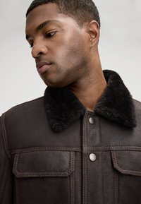 Brązowa skórzana kurtka z czarnym futrzanym kołnierzem. Posiada dwie kieszenie na piersi, zapięcia na guziki oraz widoczne przeszycia.