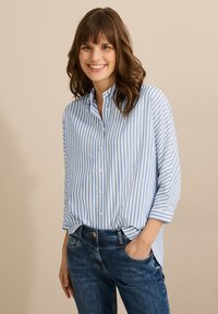 Camisa azul claro de botones con rayas verticales blancas, con un cuello redondeado y mangas arremangadas, combinada con jeans de mezclilla.
