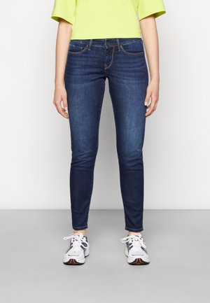 Jeans ajustados de mezclilla elástica en azul oscuro, con un diseño de cinco bolsillos, cremallera y costuras en contraste. Combinados con zapatillas blancas.